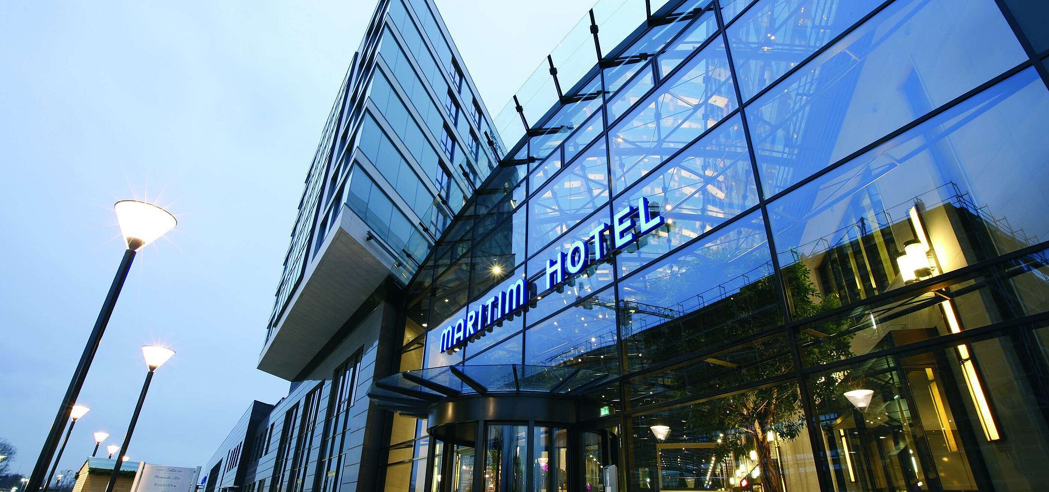 Modern exterior view of the Maritim Hotel Düsseldorf with glass facade and illuminated hotel logo at dusk.