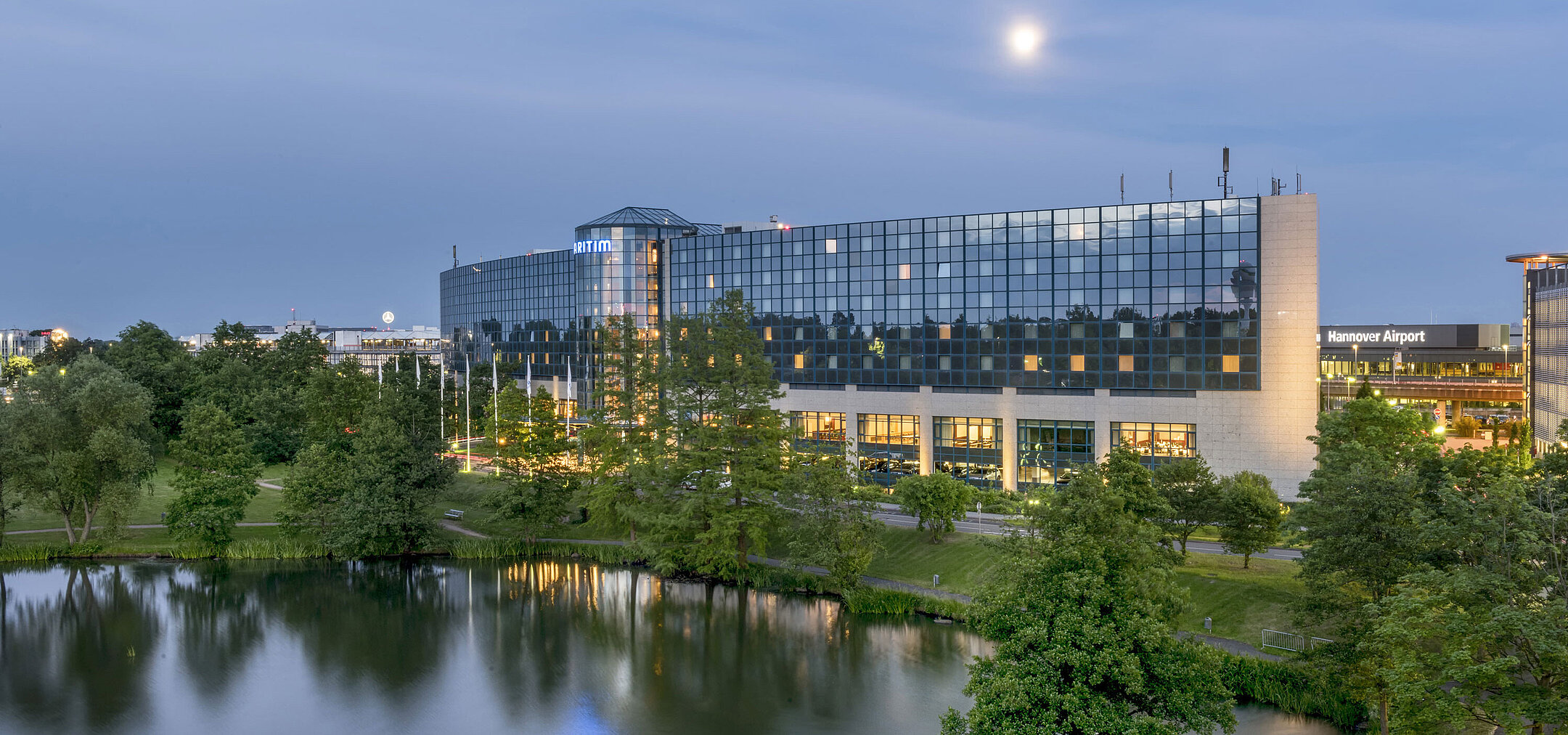 Illuminated Maritim Hotel Hannover with a glass facade and lake reflection at dusk.