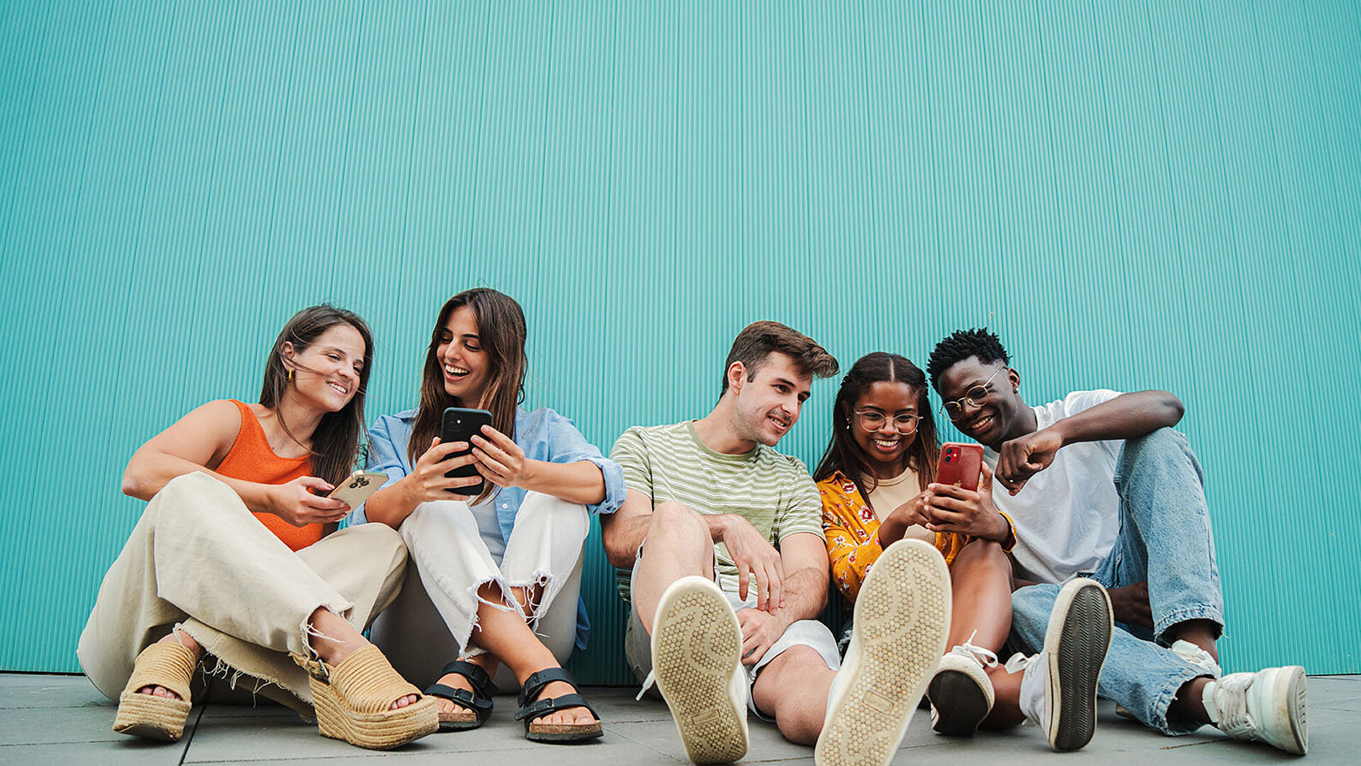 Junge Leute sitzen vor einer blauen Wand, lachen und schauen gemeinsam auf ihre Smartphones