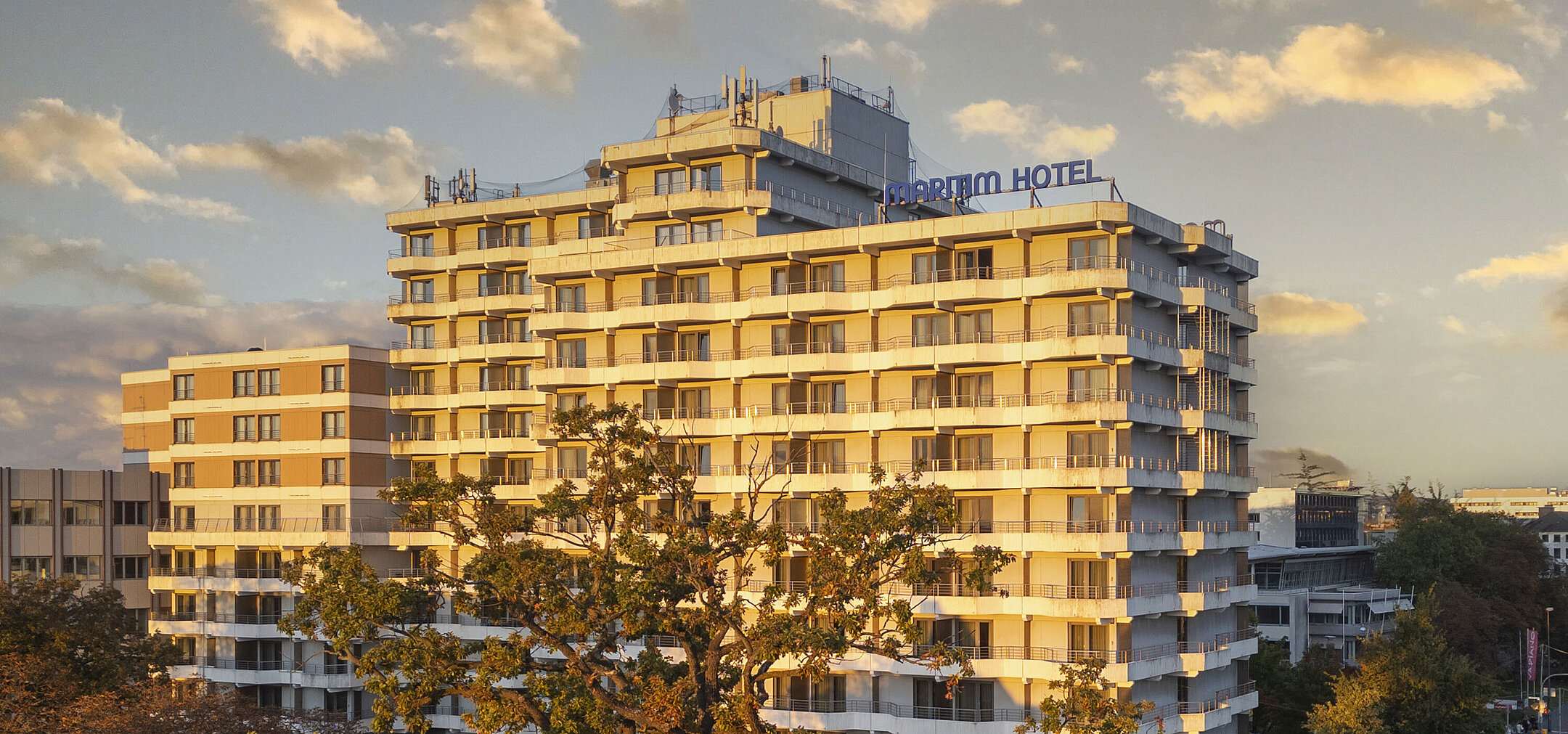 Exterior view of the Maritim Hotel Darmstadt at sunset with modern architecture and trees in the foreground.