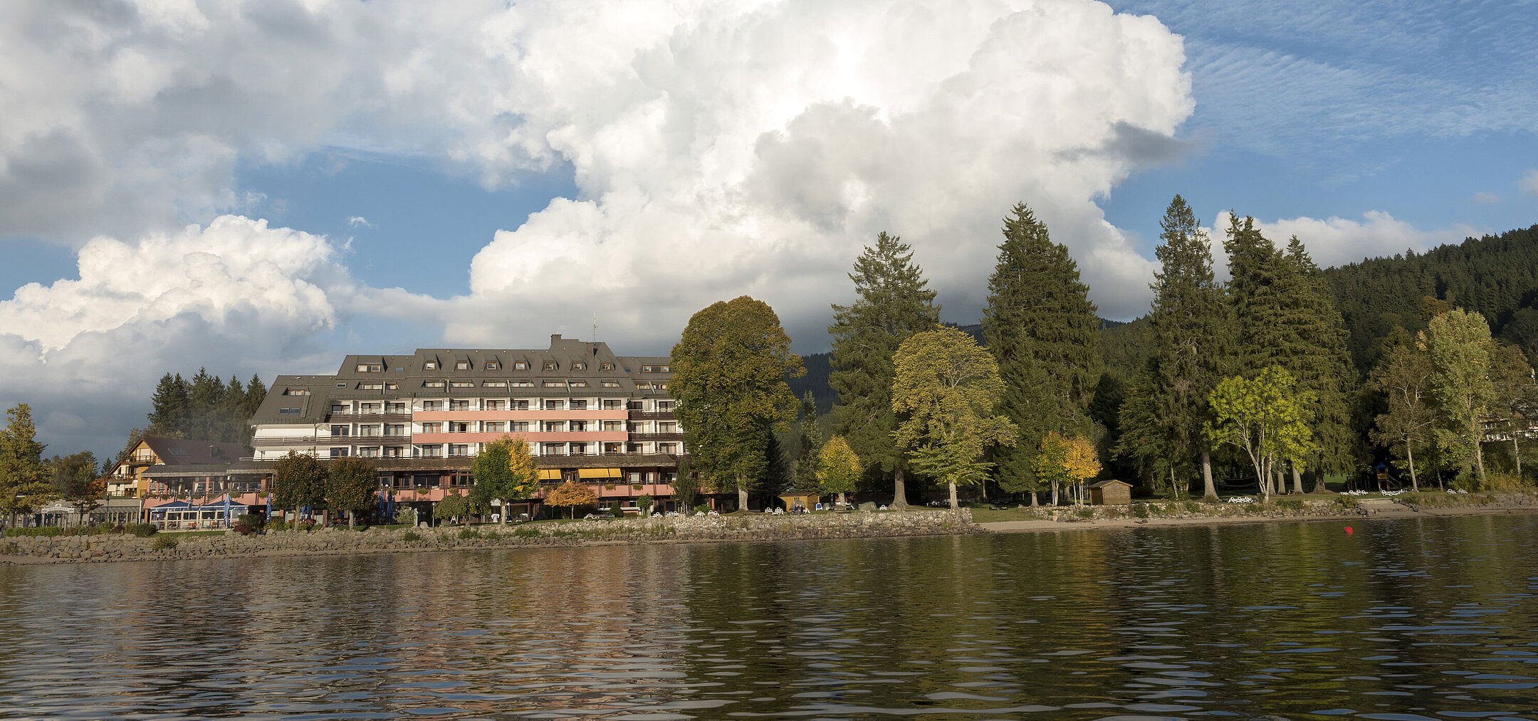 Exterior view of Maritim Hotel Titisee with trees and lakeside under blue sky and clouds