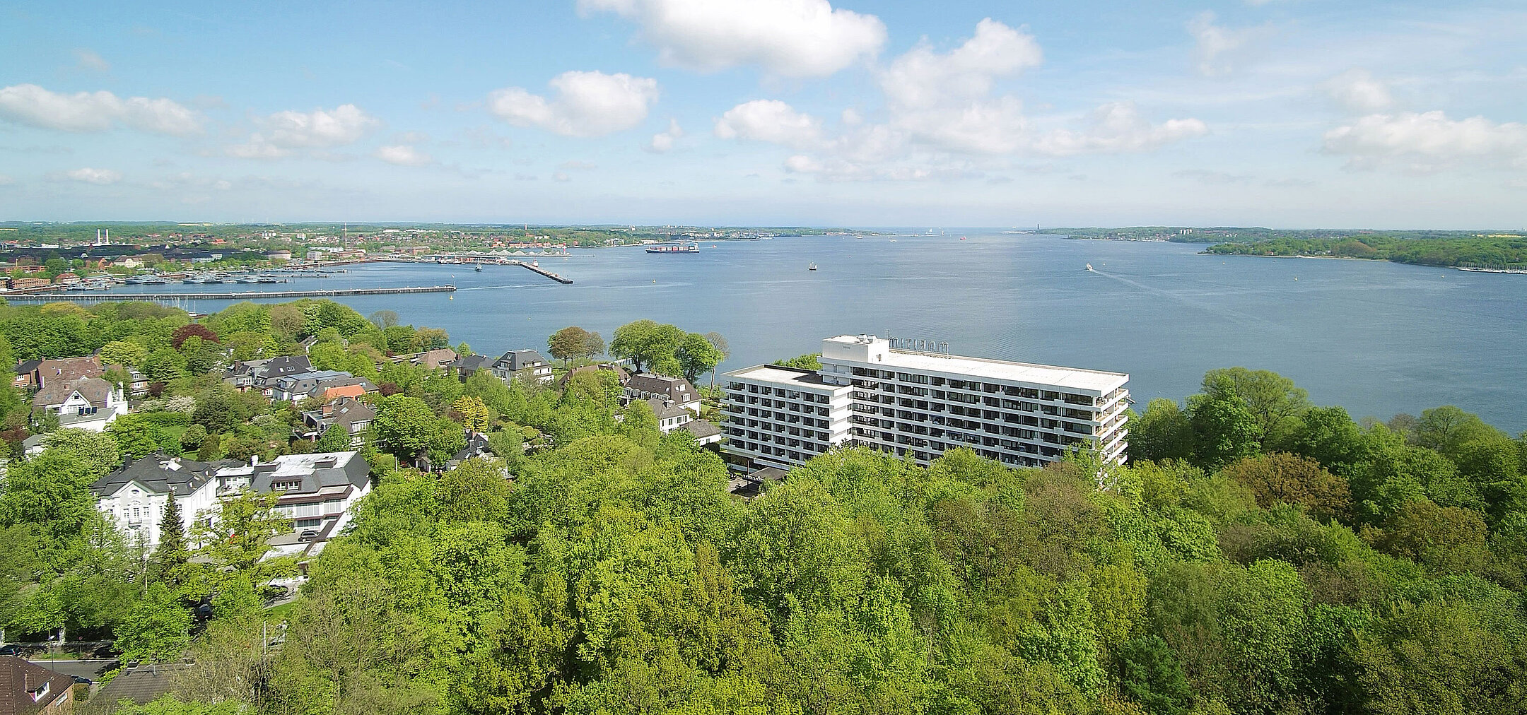Aerial view of the Maritim Hotel Kiel, surrounded by green countryside and the Kiel Fjord, under a bright blue sky.